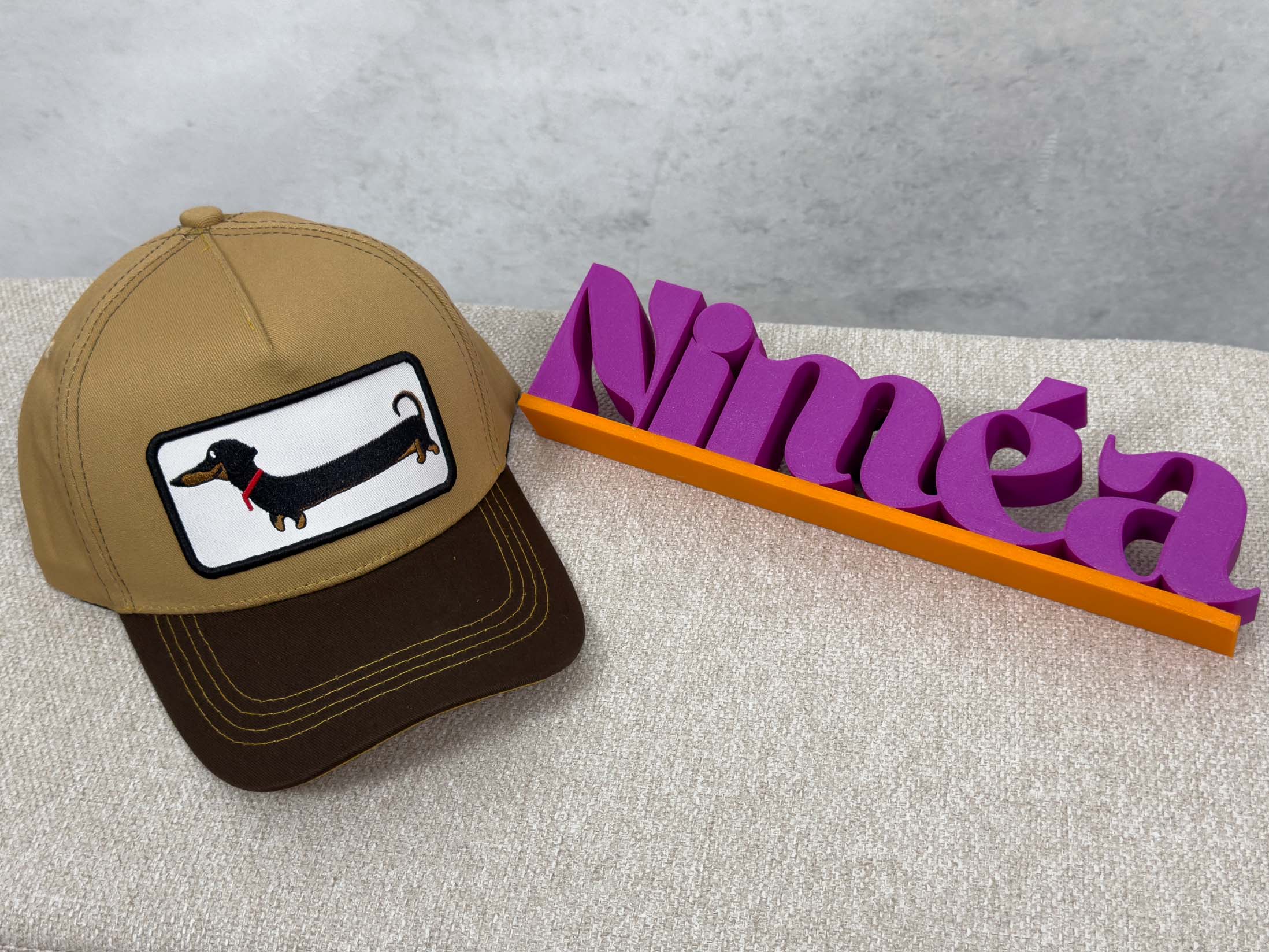 Beige Trucker Cap mit dunkelbraunem Schirm und rechteckigem Patch mit schwarzem Dackel und rotem Halsband.