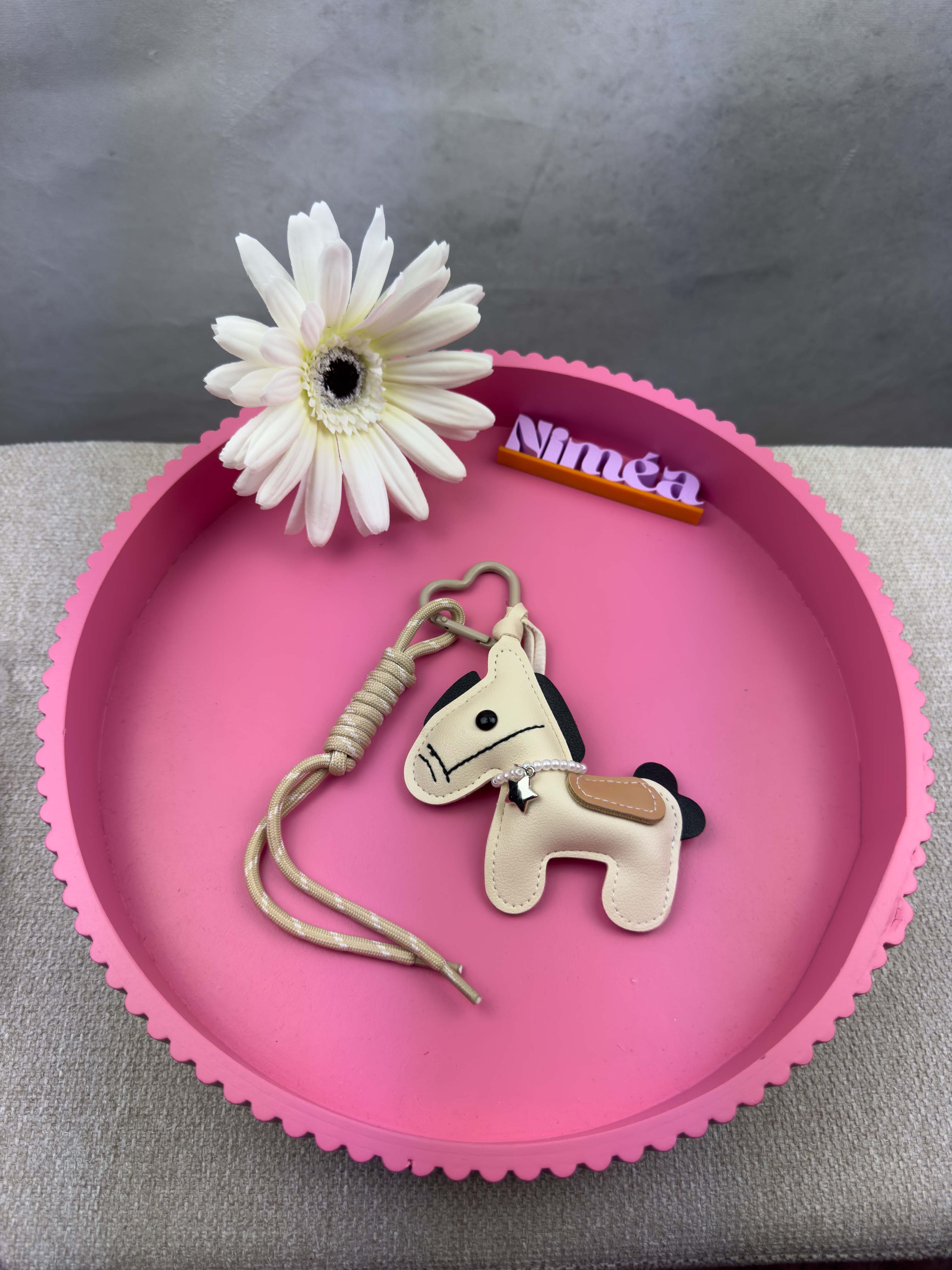 Ein kleiner beiger Pferde Schluesselanhaenger aus Leder mit beiger Kordel und Herz Karabiner liegt auf einem runden rosa Tablett neben einer weissen Blume und einem Namensschild mit der Aufschrift Nina