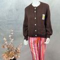 Locker geschnittener brauner Strickcardigan mit Rundhalsausschnitt, weisser Herz Knopfleiste und aufgenaehtem Smiley Patch auf Brusthoehe