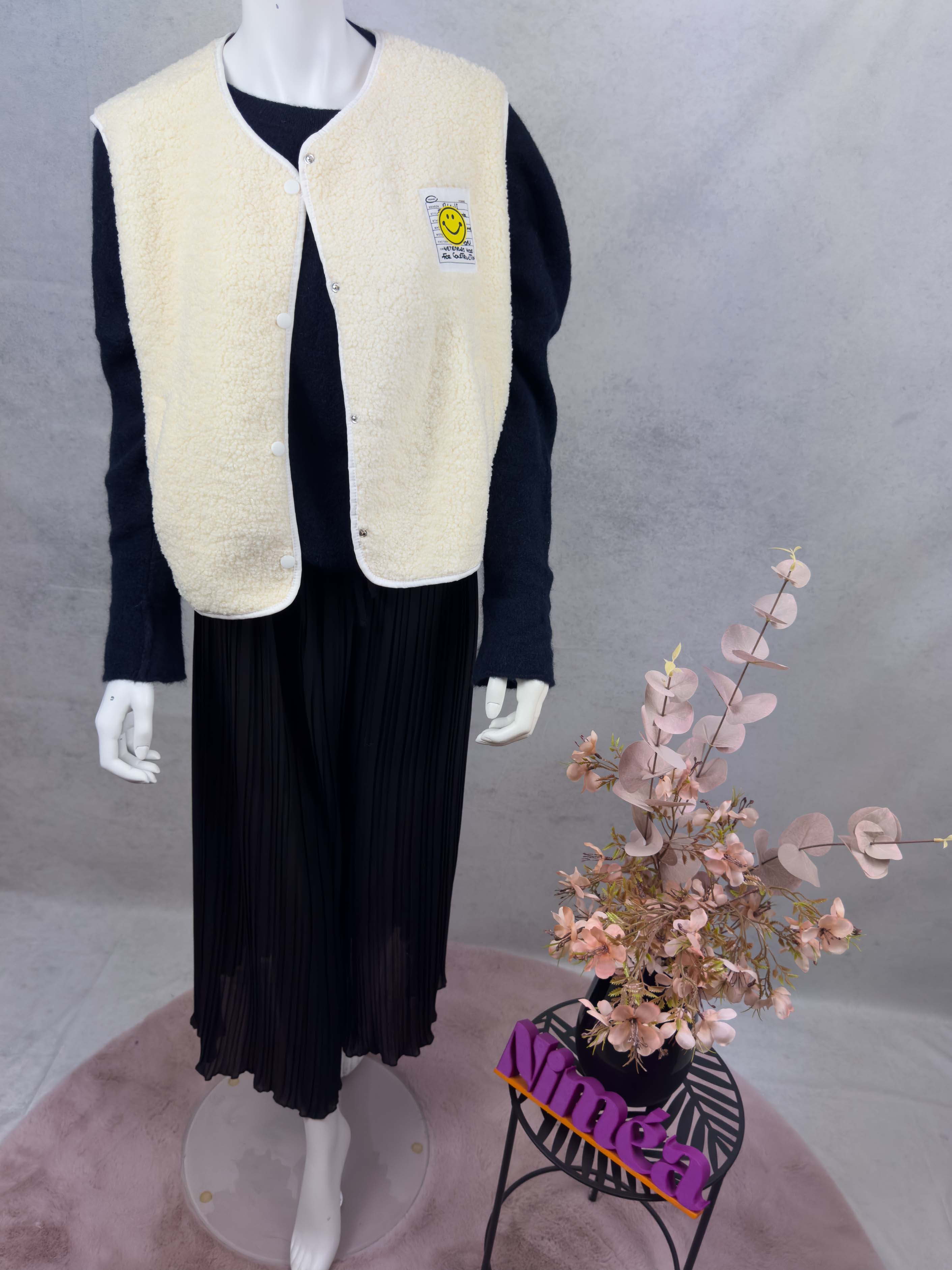 Cremefarbene Teddy-Weste mit flauschiger Oberflaeche, Druckknoepfen und Smiley-Patch auf der Brust.