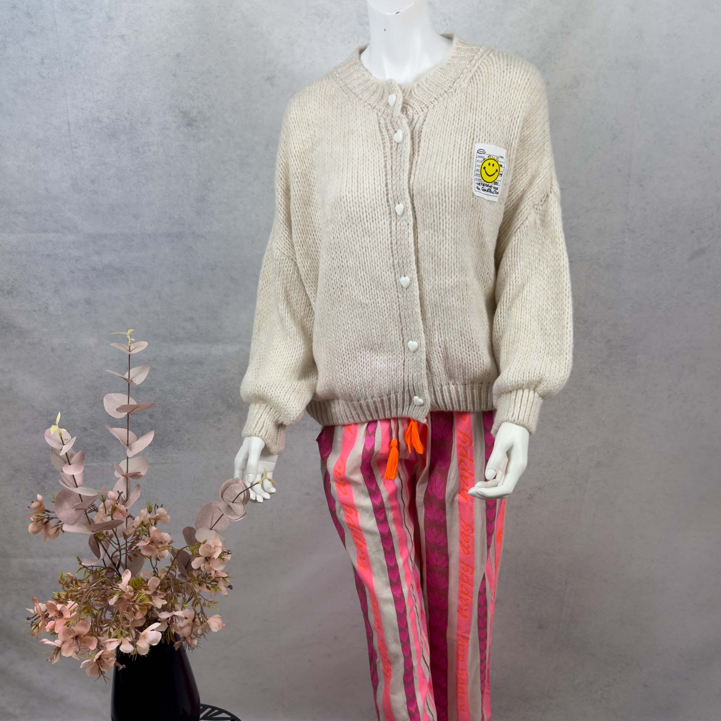 Locker geschnittener cremefarbener Strickcardigan mit Rundhalsausschnitt, weisser Herz Knopfleiste und aufgenaehtem Smiley Patch auf Brusthoehe