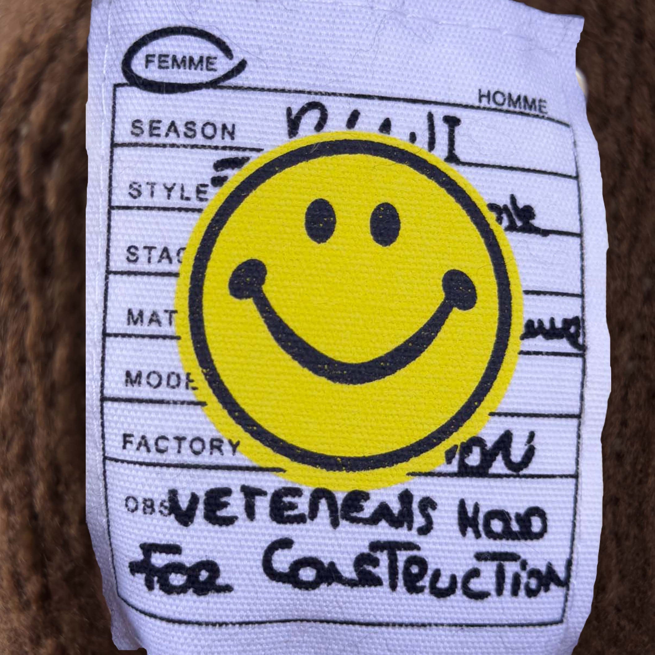 Nahaufnahme des aufgenaehten Smiley Labels mit Schriftzug auf braunem Strick mit sichtbarer grober Strickstruktur