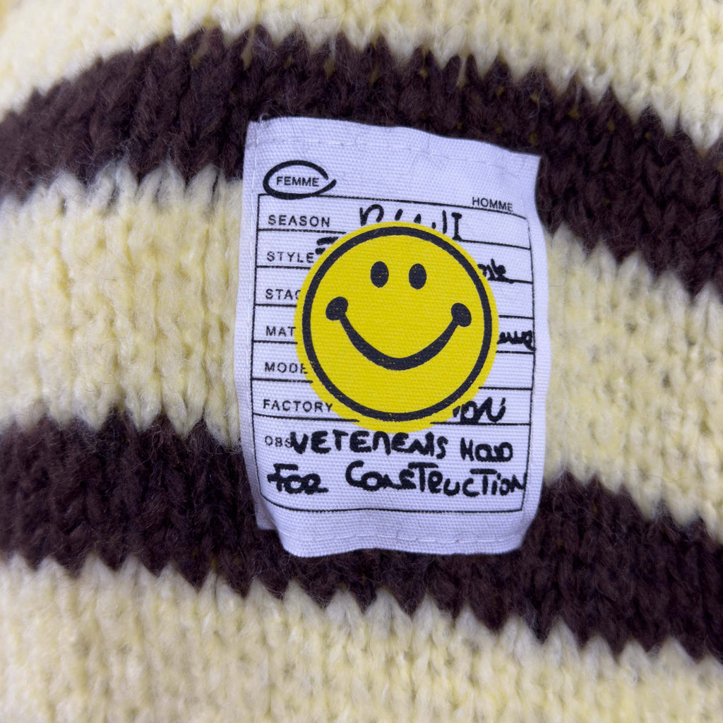Nahaufnahme des aufgenaehten Smiley Labels auf gelb braun gestreiftem Strick mit sichtbarer Strickstruktur