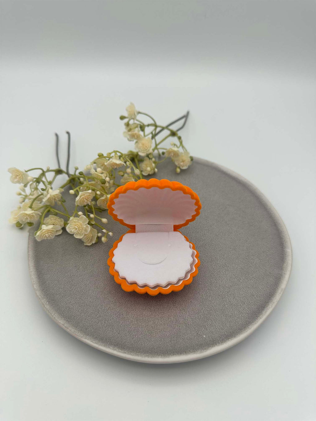 Orangefarbenes Schmuckkaestchen in Muschelform, geoeffnet, mit heller gepolsterter Innenflaeche fuer Ringe oder Ohrringe.