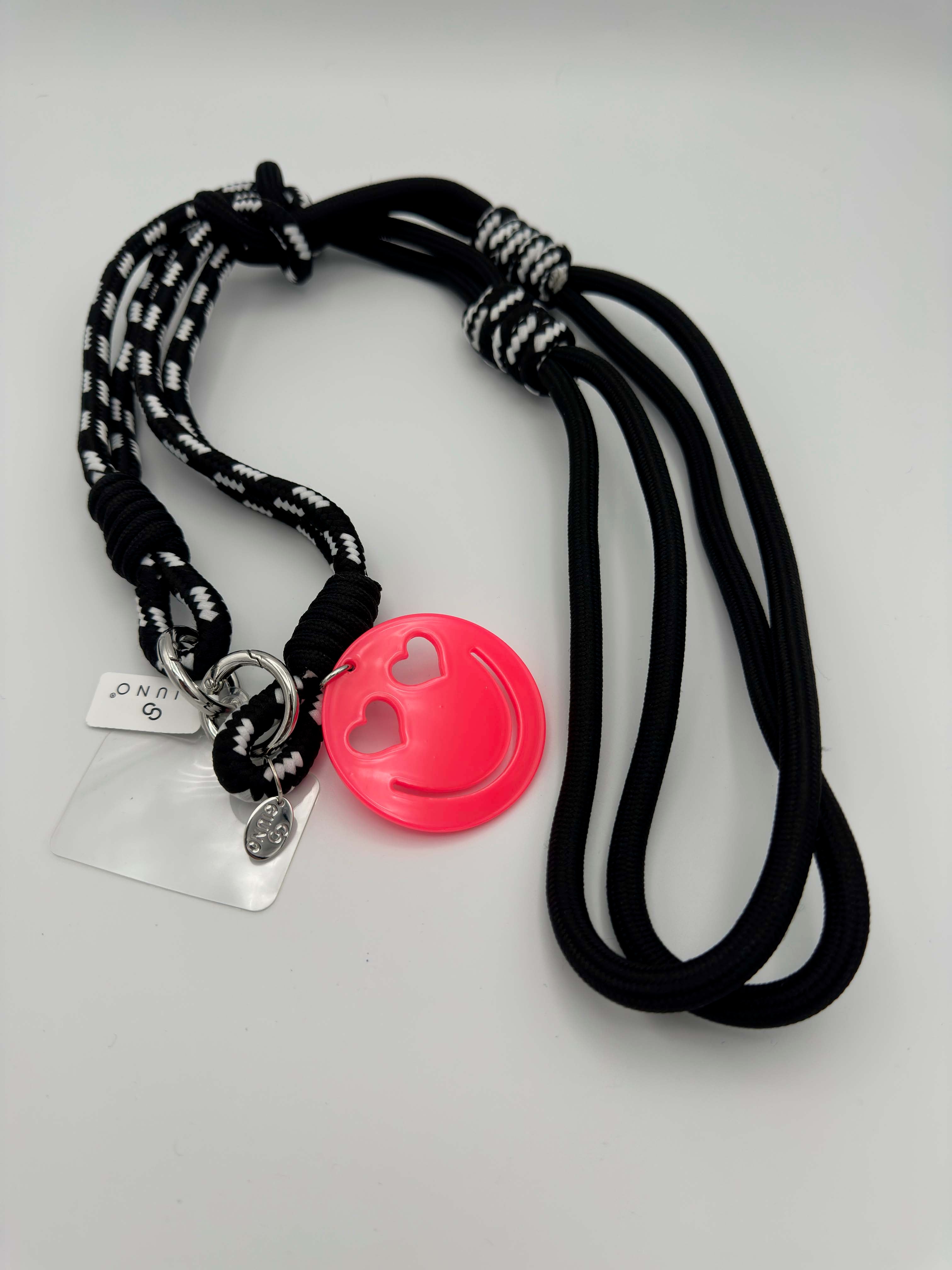 Schwarze Handykette mit schwarz-weiß gemustertem Band und neonpinkem Smiley-Anhänger mit Herzaugen.