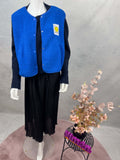 Mannequin traegt eine blaue Teddy-Weste mit gelbem Smiley-Patch auf der linken Brust, schwarzem Oberteil und schwarzem Plisseerock; dekoriert neben einem kleinen Tisch mit Blumen.