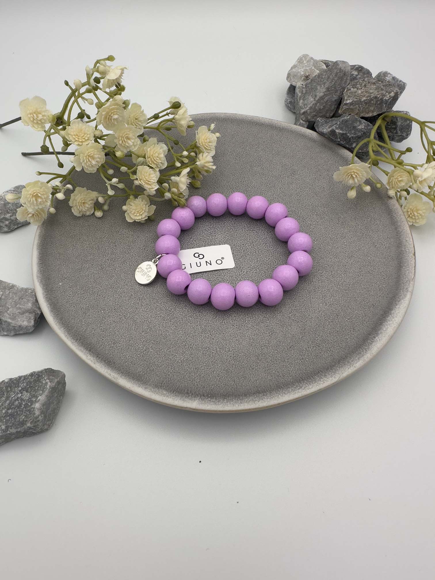 Elastisches Armband aus lilafarbenen Holzperlen mit rundem silbernen Anhänger auf hellem Hintergrund.