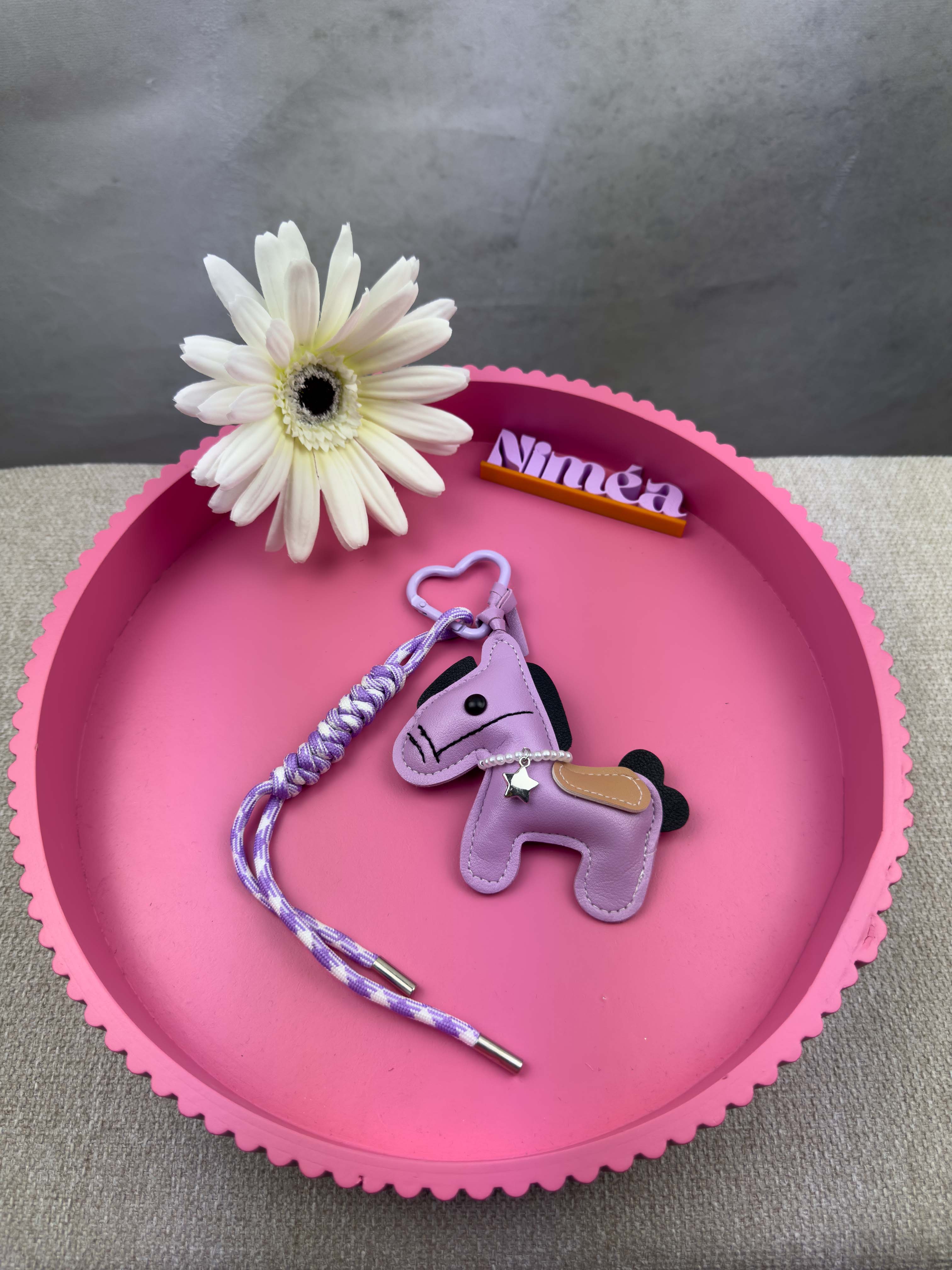 Ein kleiner lila Pferde Schluesselanhaenger aus Leder mit geflochtener lila Kordel und Herz Karabiner liegt auf einem runden rosa Tablett neben einer weissen Blume und einem Namensschild mit der Aufschrift Nina