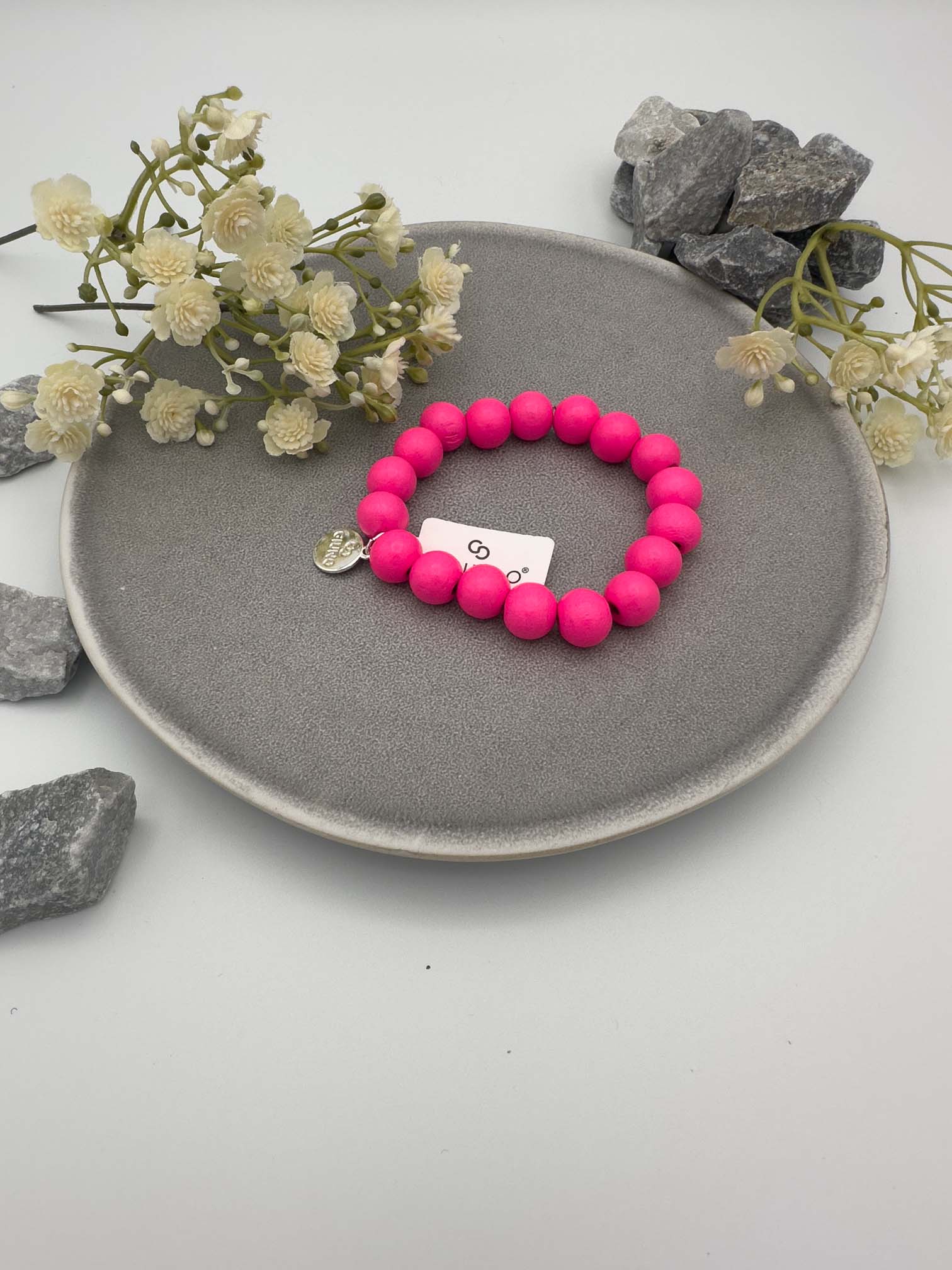Elastisches Armband aus pinkfarbenen Holzperlen mit rundem silbernen Anhänger auf hellem Hintergrund.