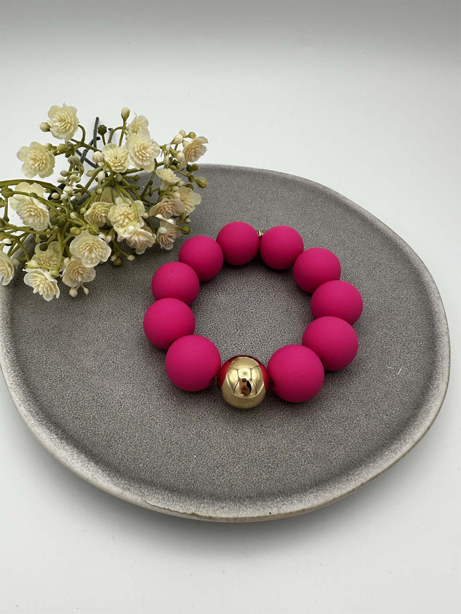 Elastisches Armband mit matten pinken Perlen und goldfarbener Kugel dekorativ auf grauem Teller arrangiert