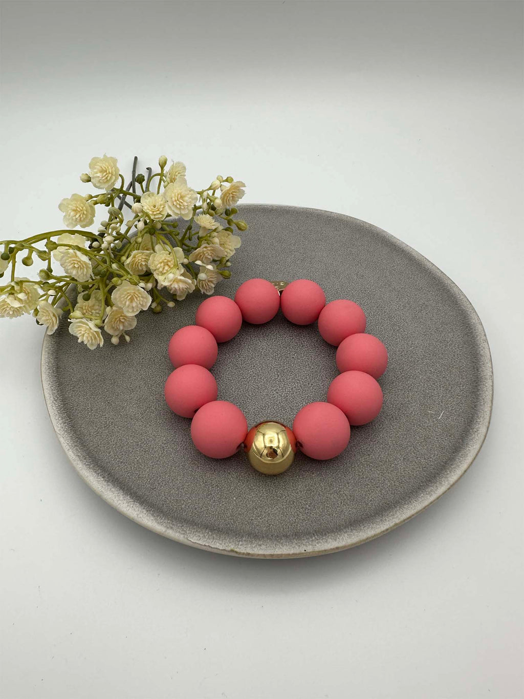 Elastisches Armband mit matten rosa Perlen und goldfarbener Kugel auf grauem Teller mit dekorativen Blumen