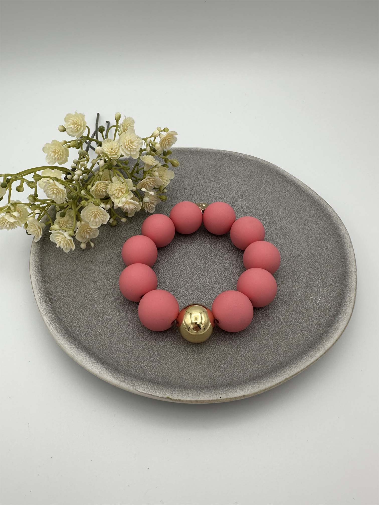 Elastisches Armband mit matten rosa Perlen und goldfarbener Kugel auf grauem Teller mit dekorativen Blumen