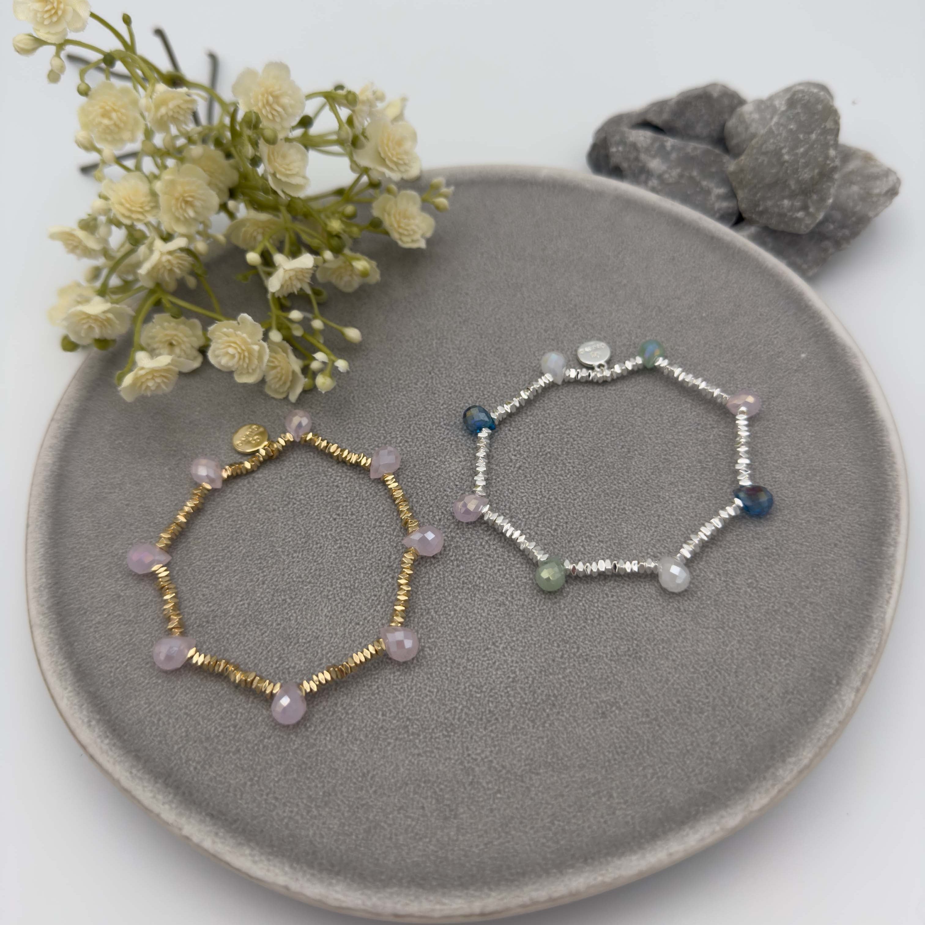 Zwei elastische Armbaender, eines goldfarben mit rosefarbenen facettierten Glasperlen, eines silberfarben mit facettierten Perlen in Blau, Gruen und Grau.